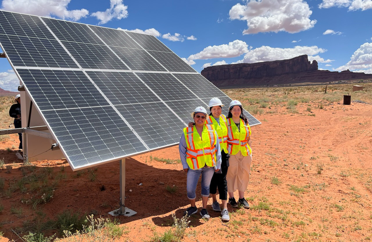 Three Native American woman in front of a solar panel in the desert.