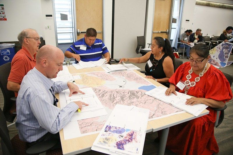 Five people seated around a table looking at a map and documents.