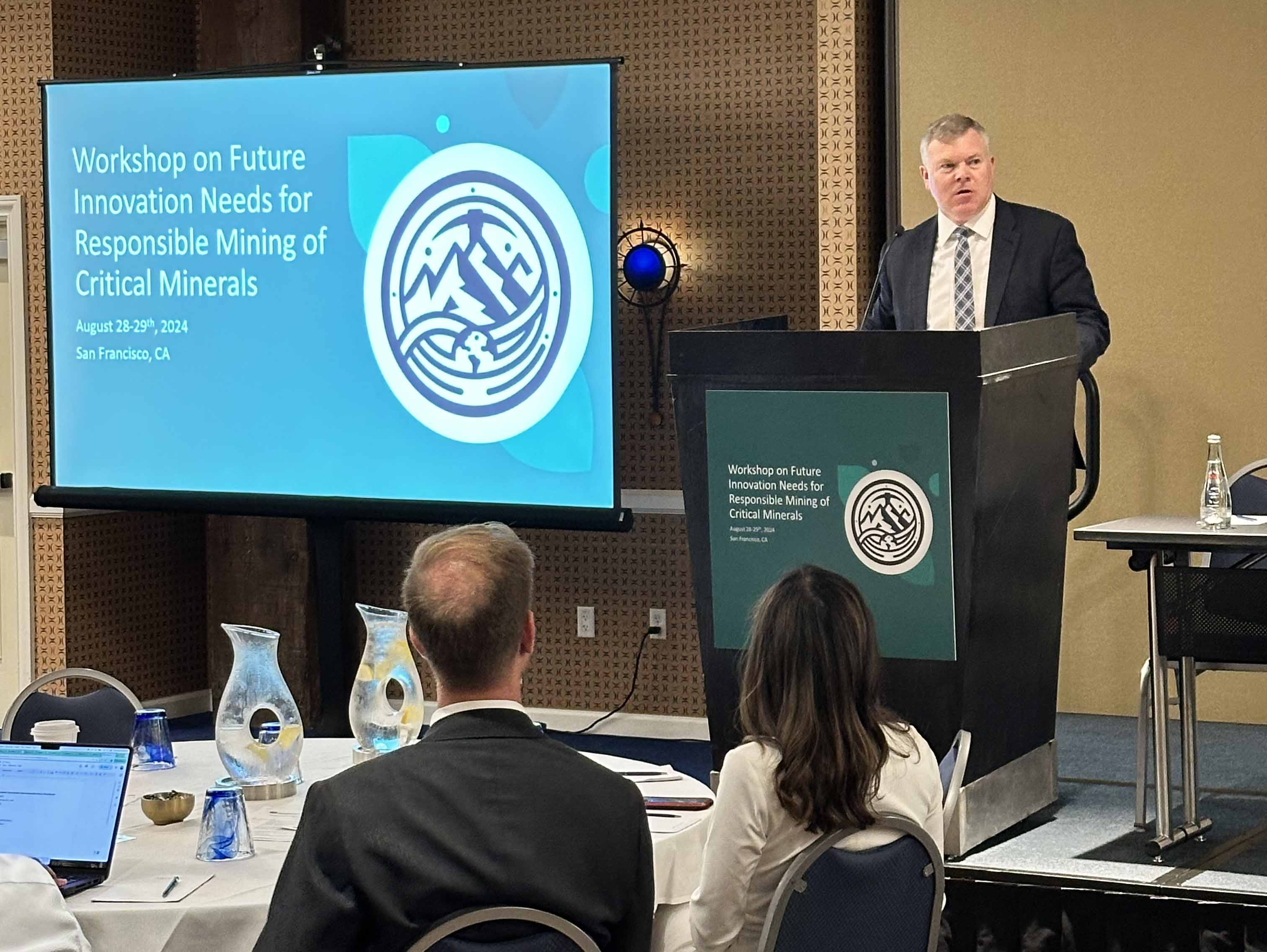 A person speaking behind a podium next to a screen with a slide titled "Workshop on Future Innovation Needs for Responsible Mining of Critical Materials"