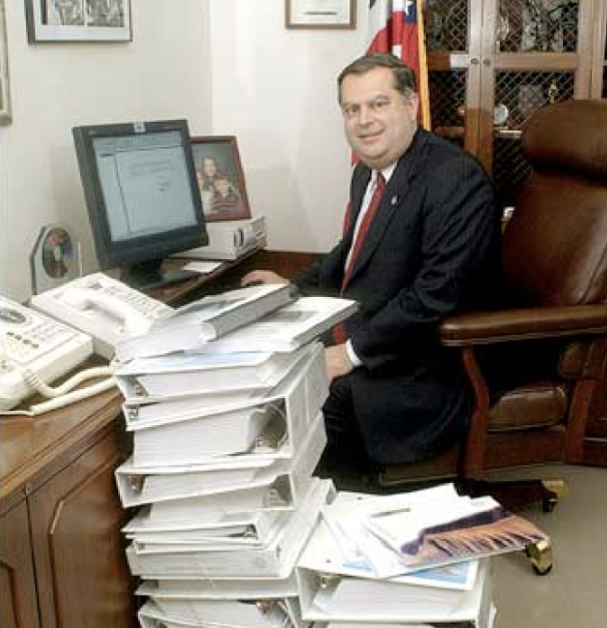 Secretary Abraham seated at his office desk, surrounded by a stack of binders almost as tall as him. He sits in front of a late 90's style computer monitor.