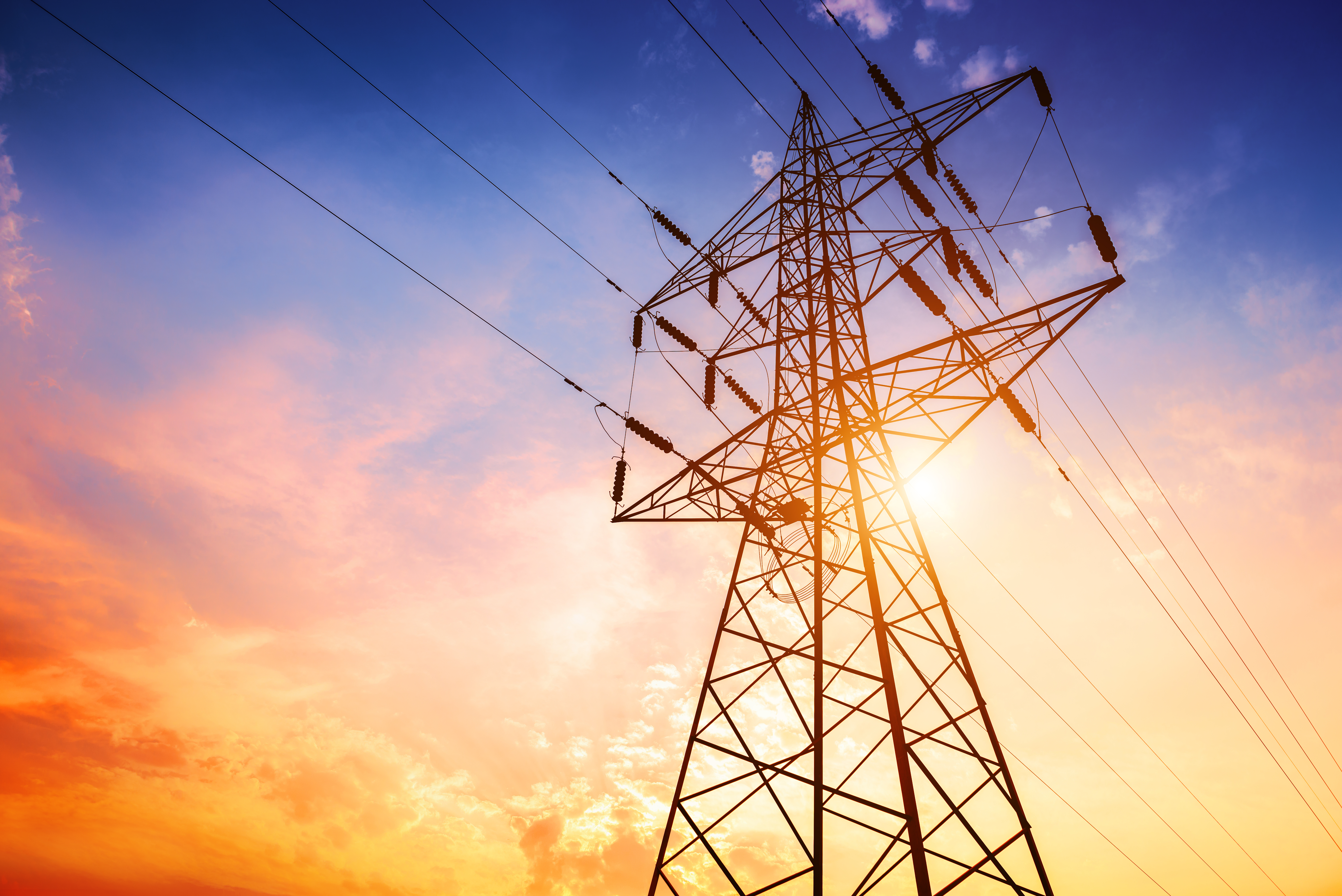 Photo looking up at a large power transmission line, with the sun behind it, and the sky a rich hue of blues, pinks, and oranges.