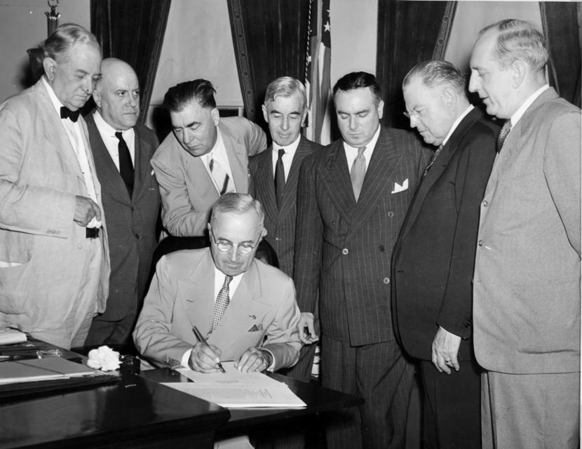 Black and white photo of President Eisenhower signing the Atomic Energy Act in 1954. He sits at a table with a group of men around him in suits.