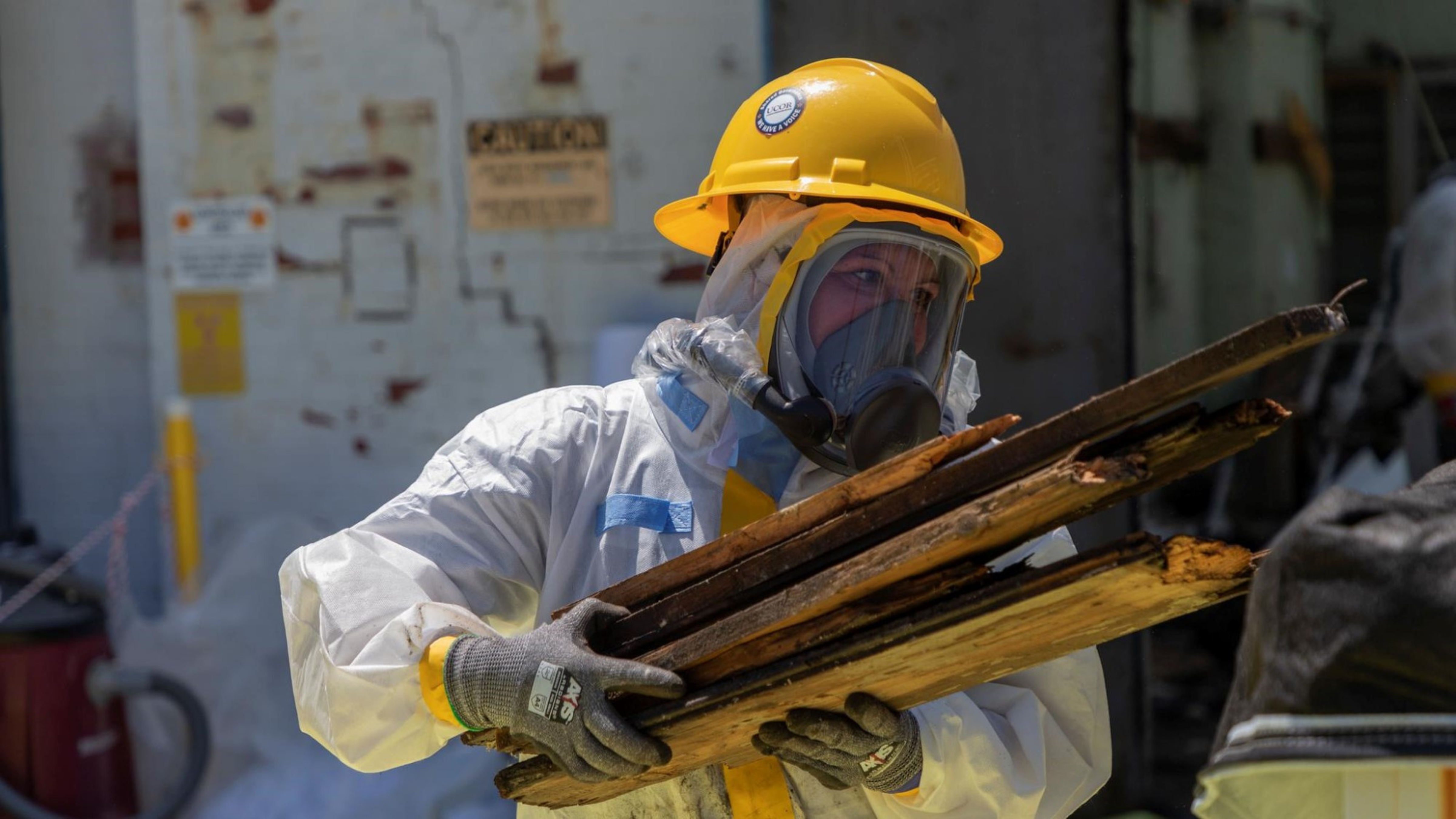 A person in protective gear carries wood in their arms. 