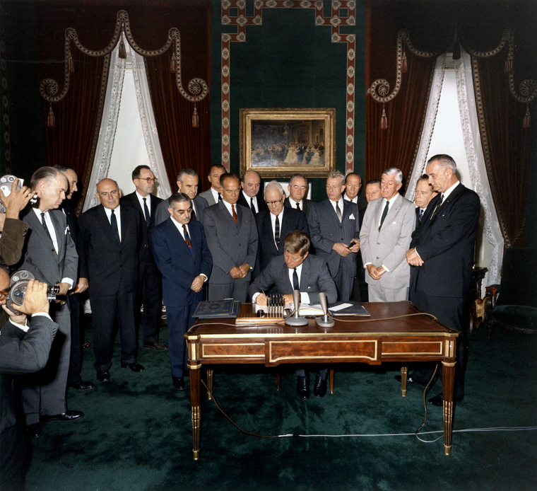 Photo of President JFK signing the Limited Nuclear Test Ban Treaty in 1963. It is a color photo; Kennedy is seated at a table signing the ban, surrounded by a group of men in suits.