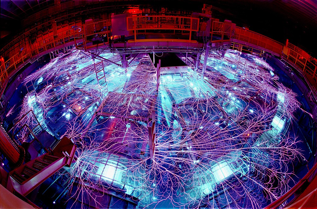 Photo of the Z Machine, part of the Pulsed Power Program, at Sandia National Lab. The room is dark and the machine is glowing blue and purple, with reddish/pink wires everwhere.