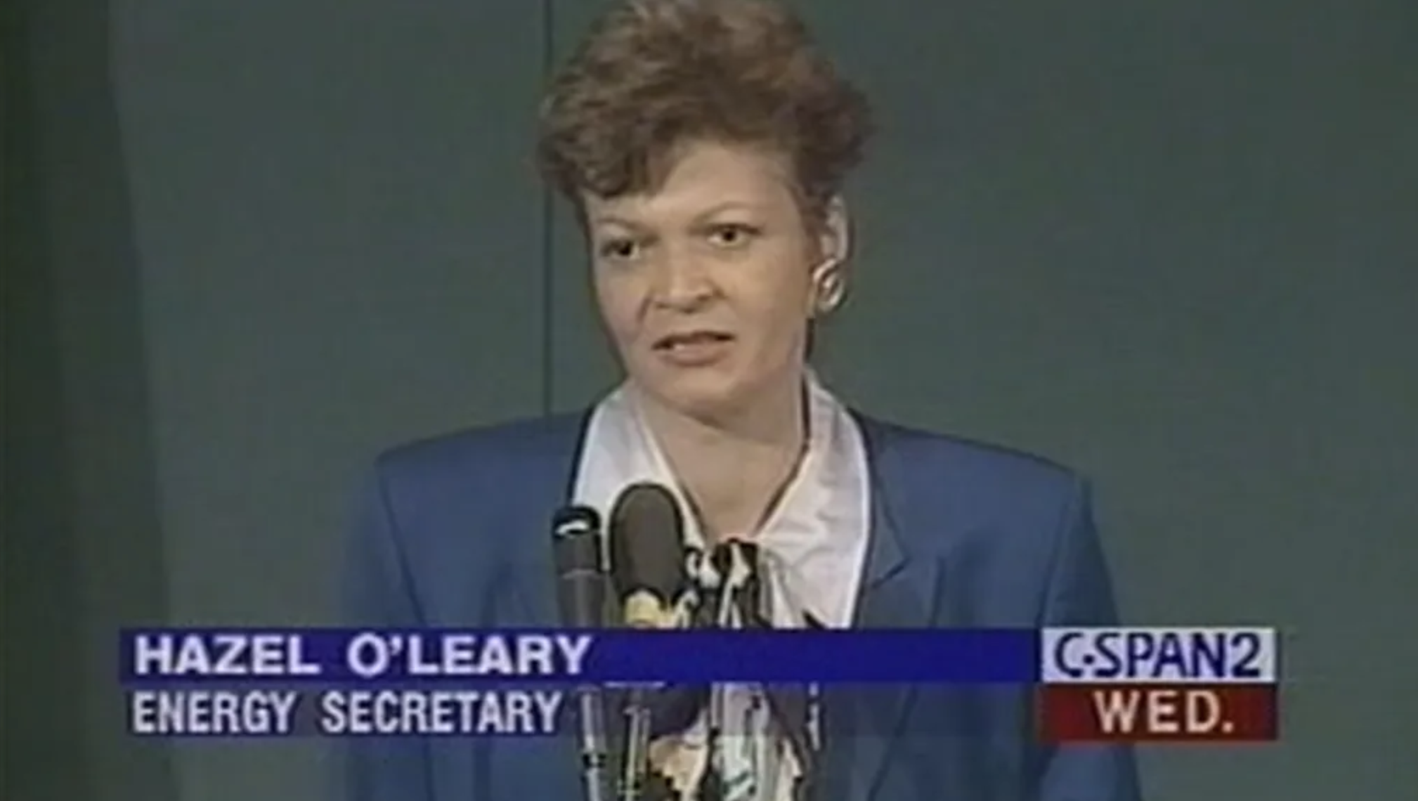 Video screengrab of original CSPAN footage as Secretary O'Leary announces the Openness Initiative. She is standing in front of a microphone where a blue suit jacket and white collared shirt.