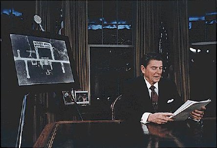 President Reagan sits at Resolute Desk in White House Oval Office, announcing the Strategy Defense Initiative. He is looking down at papers as he speaks.