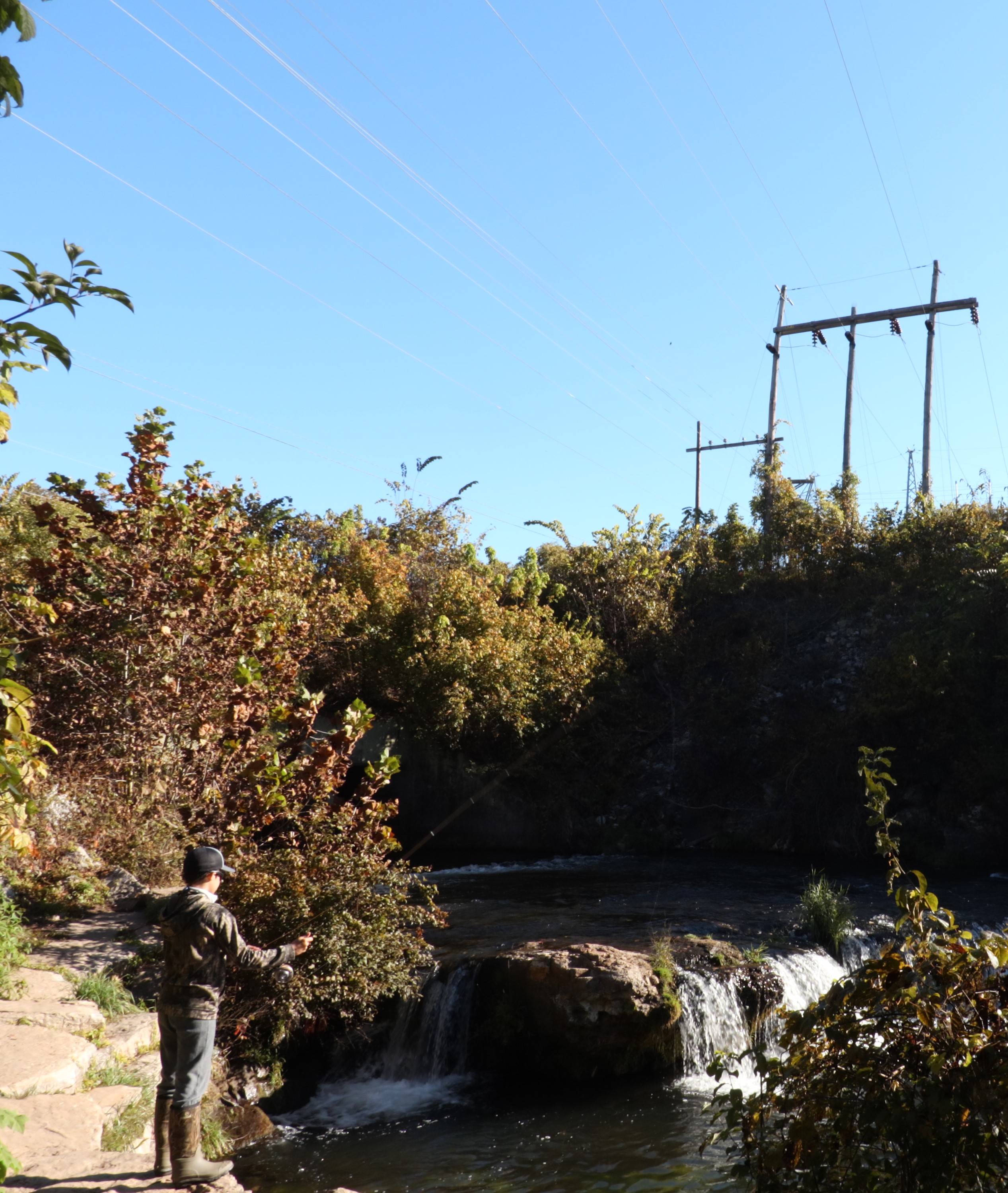 child fishing near small waterfall with transmission lines overhead