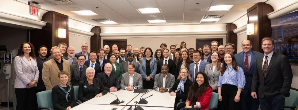 A group of 46 people in business attire pose and smile for the camera in a meeting room. 