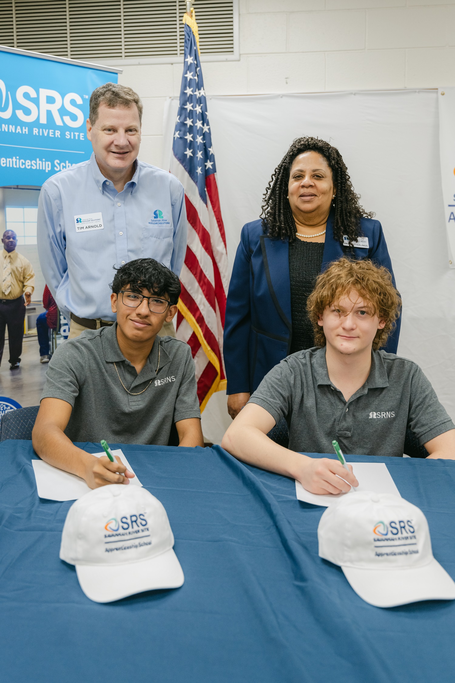 Two students sit at a table with two adults standing behind, they all pose for a picture