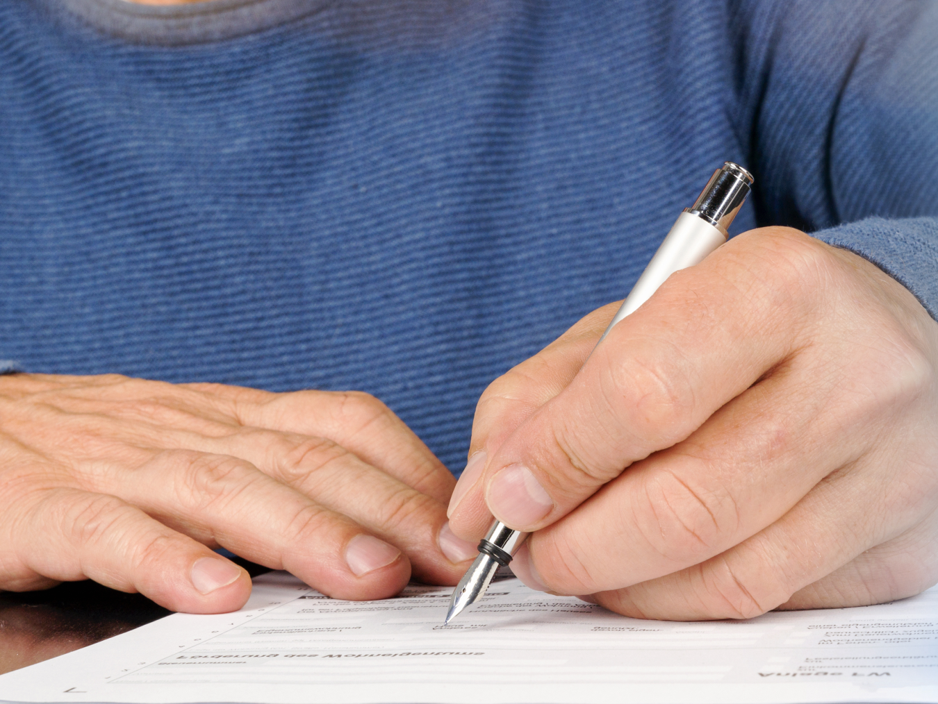 Person in blue shirt, writing on piece of paper with a white and silver tipped pen