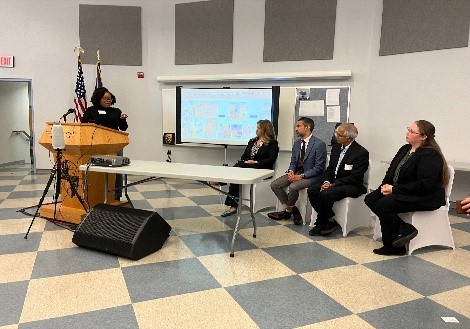 A woman standing at a podium speaks to a panel of four people sitting at the front of a large room