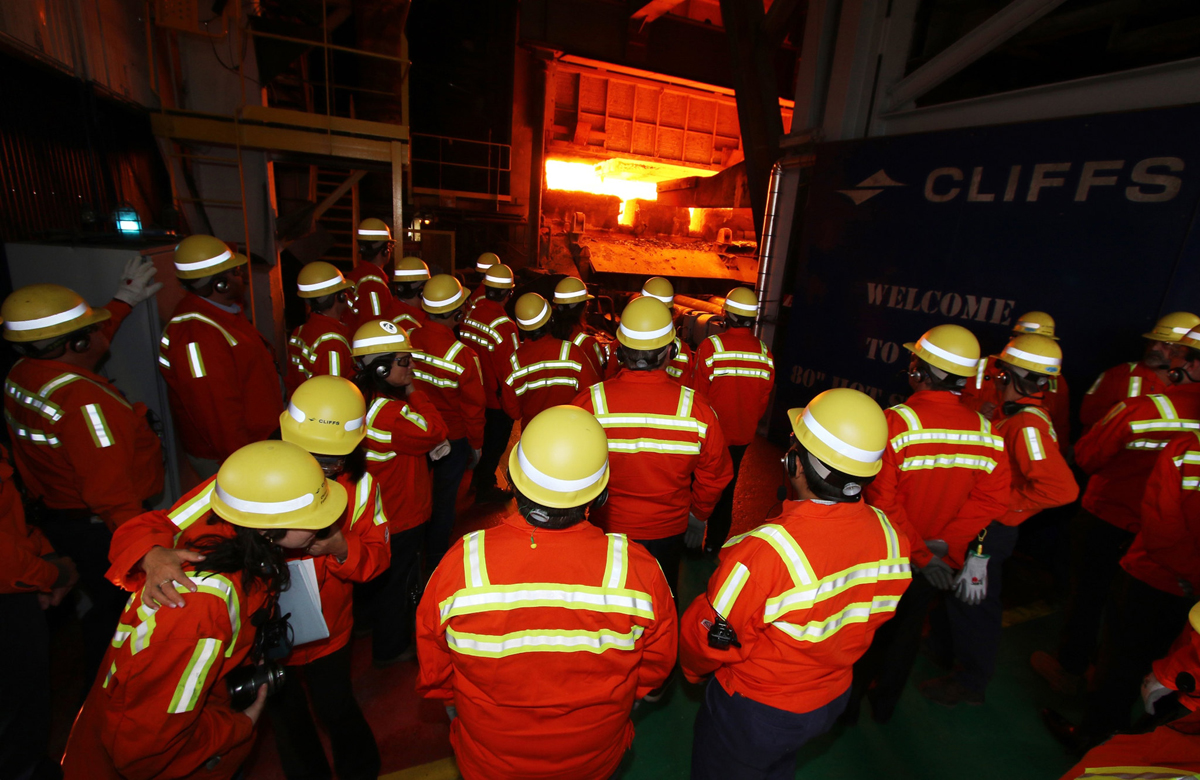 A group of researchers in hard hats and safety vests stand in front of a furnace.