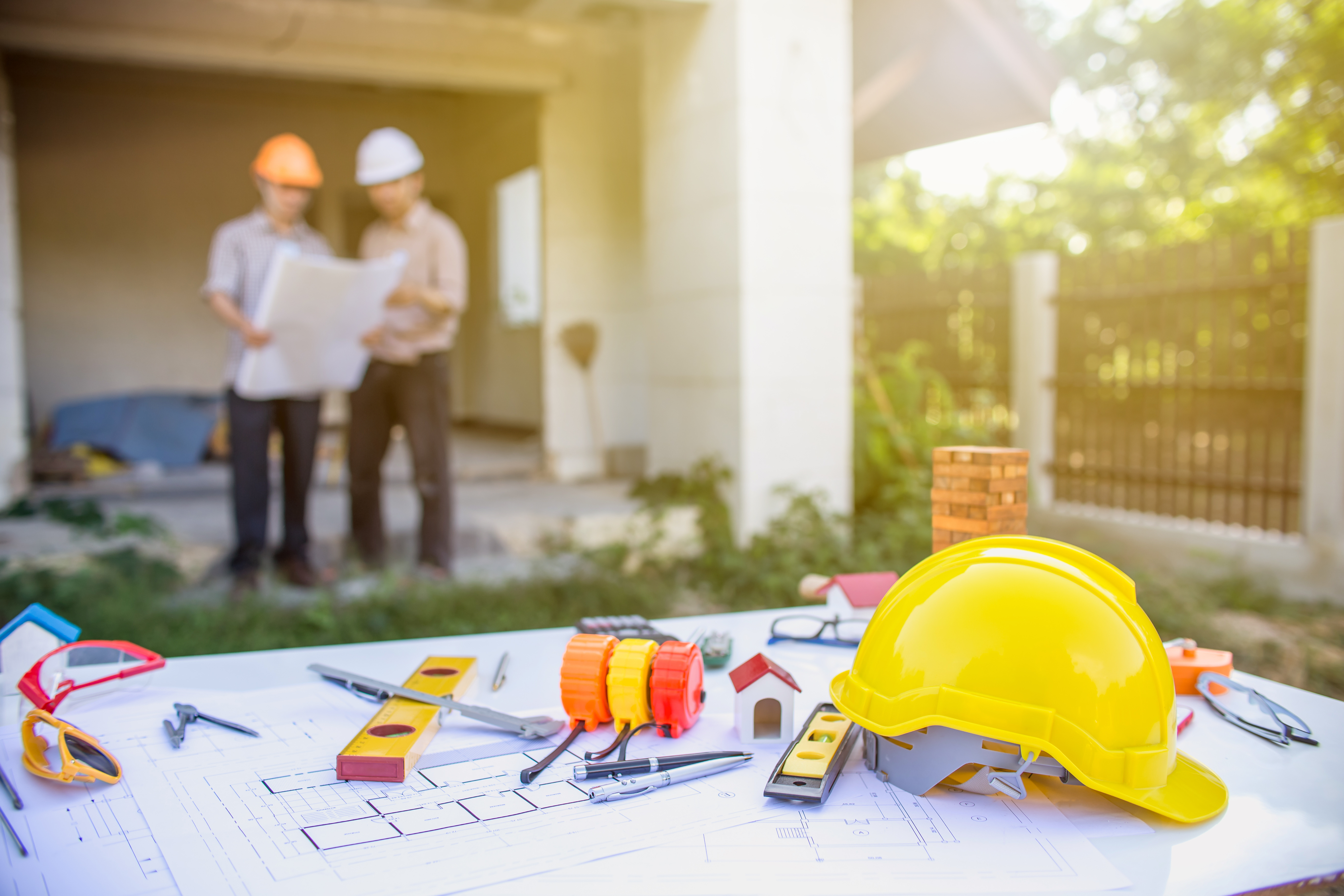 A table with various tools in the foreground, and two people in hard hats holding blueprints in the background.