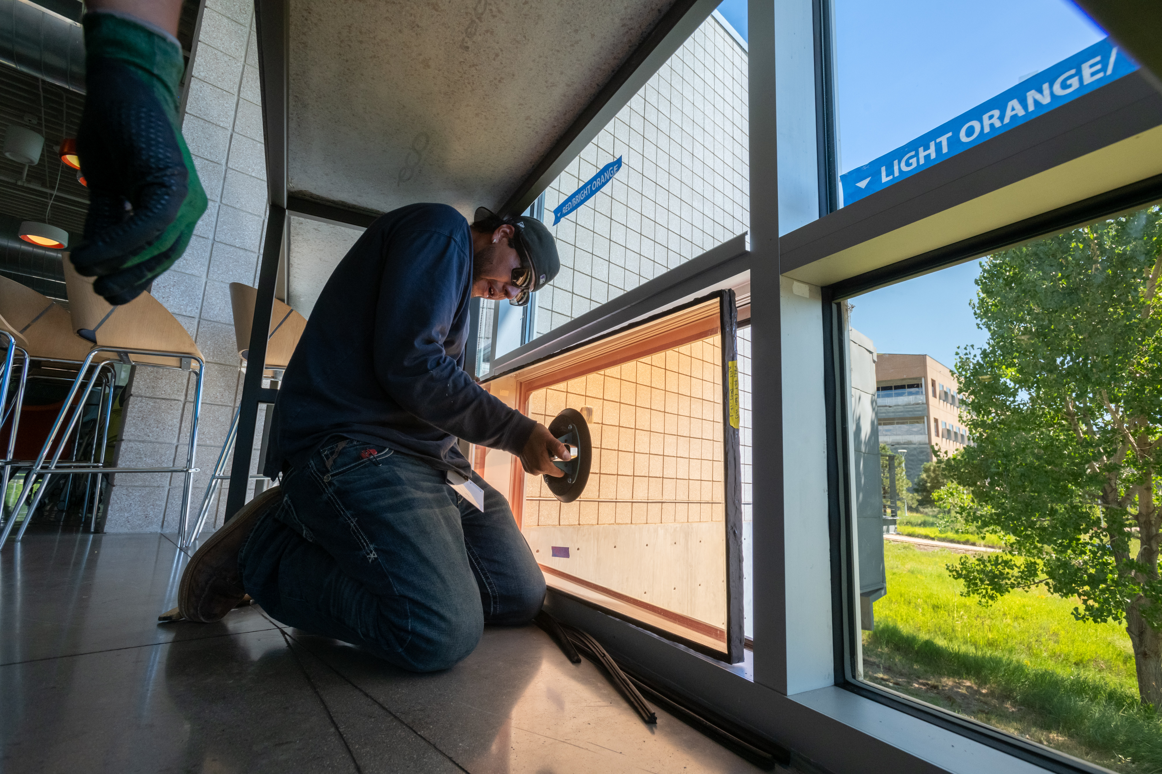 A man crouched near a lower panel of a large set of windows in a commercial building, replacing the glass in the panel.