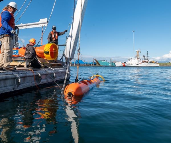 NREL researchers releasing a mooring device off a boat platform into a bay with blue sky in the background. 