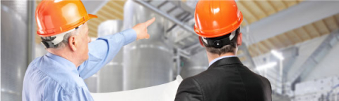 Two men in hard hats, backs facing the camera, discussing ductwork in a building.