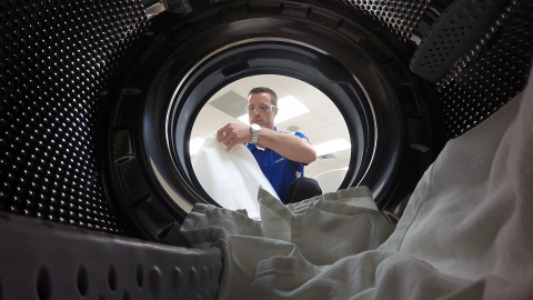 Photo from the perspective of the inside of a washing machine, showing the door open and a man with laundry beyond.