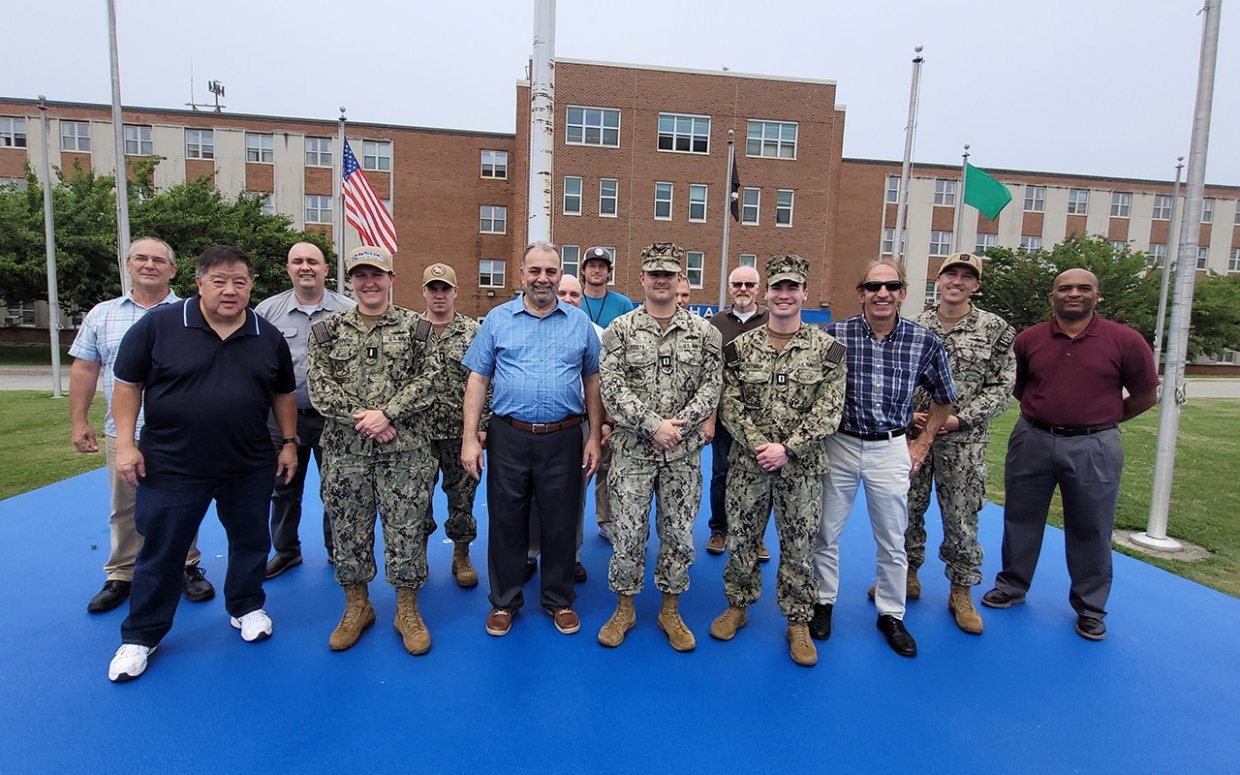 A group of people including servicemen and civilians are standing outside in front of a building smiling for the camera.