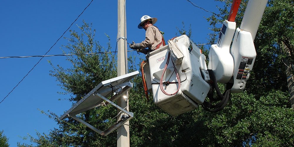 Image of a man wearing a white hard hat and standing in a lift bucket attends to a solar panel attached to a utility pole.