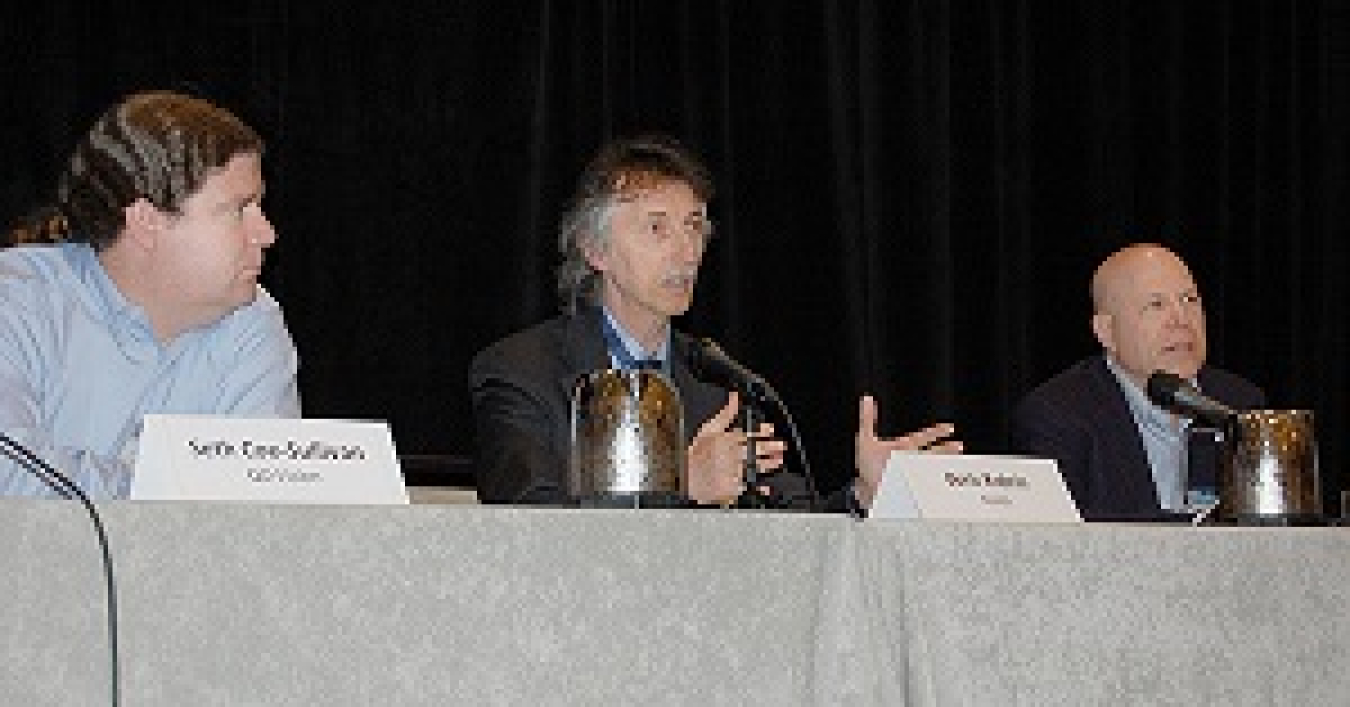 Photo of three men seated at a table, in front of microphones. The man in the middle is talking into the microphone and gesturing.