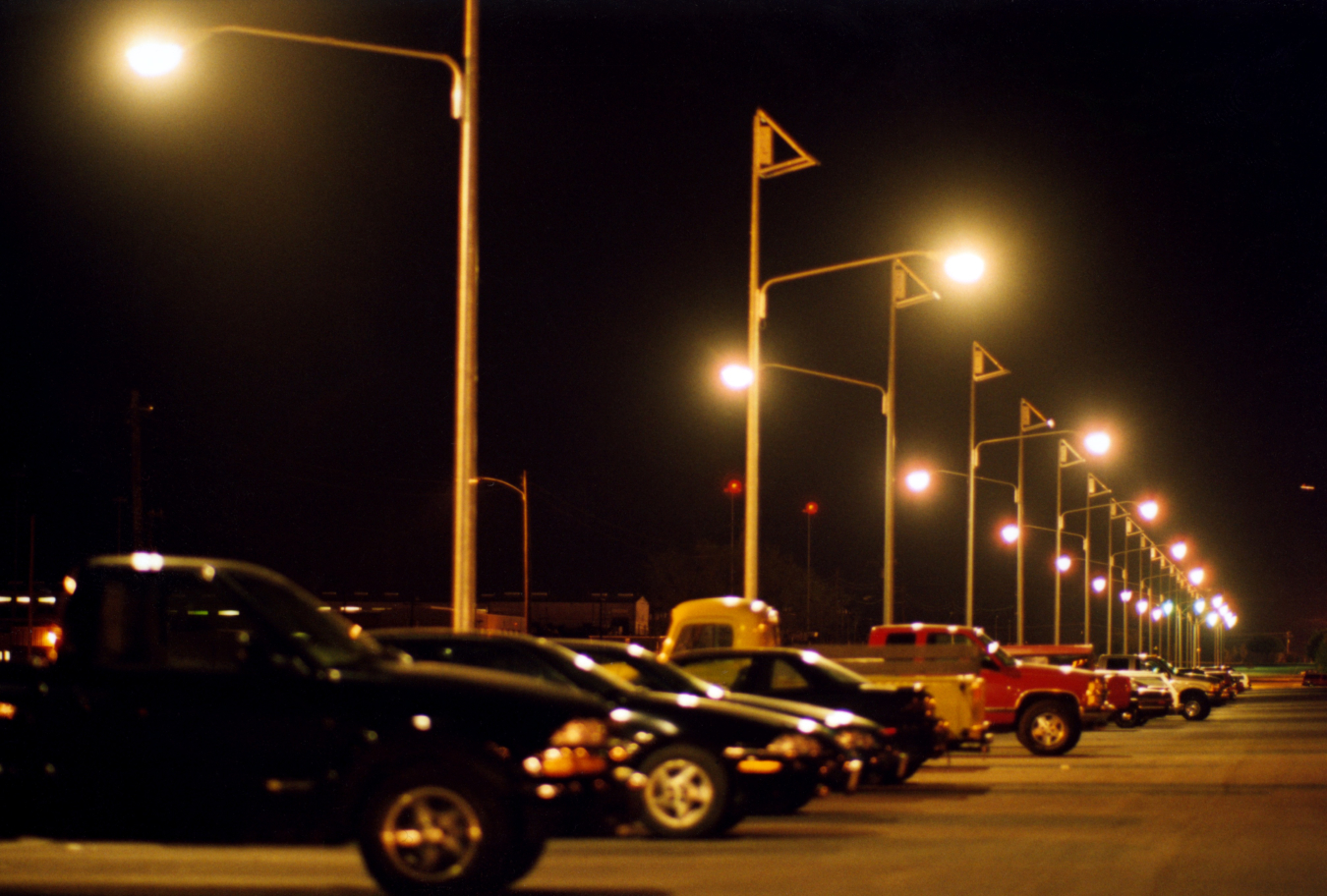 Photo of a parking lot at night with cars parked and lights lit overhead.