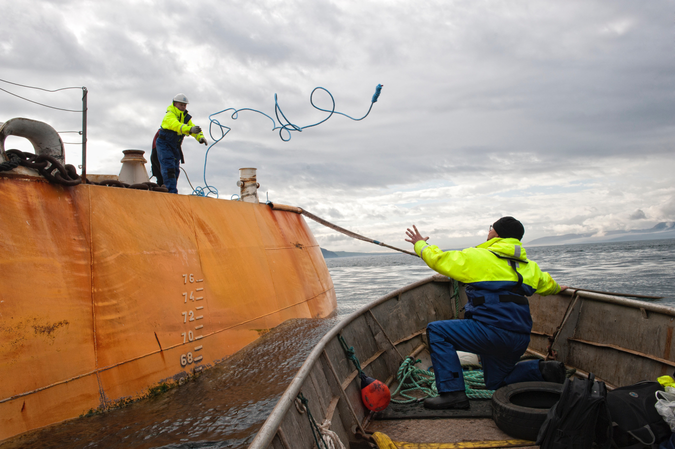 A man on a yellow vessel throws a blue rope to another man in a boat.