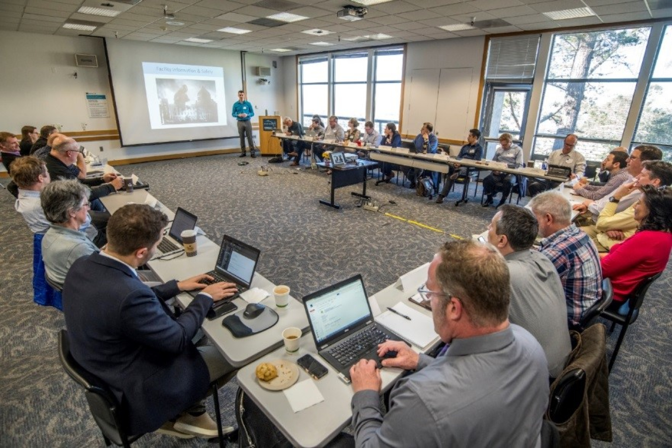 Photo of a room full of people in a meeting, seated at tables in a square configuration, with a screen showing a presentation.