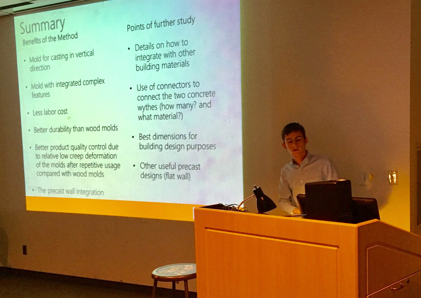 Photo of a man speaking at the head of a room near a podium, with a presentation on the large screen behind him.