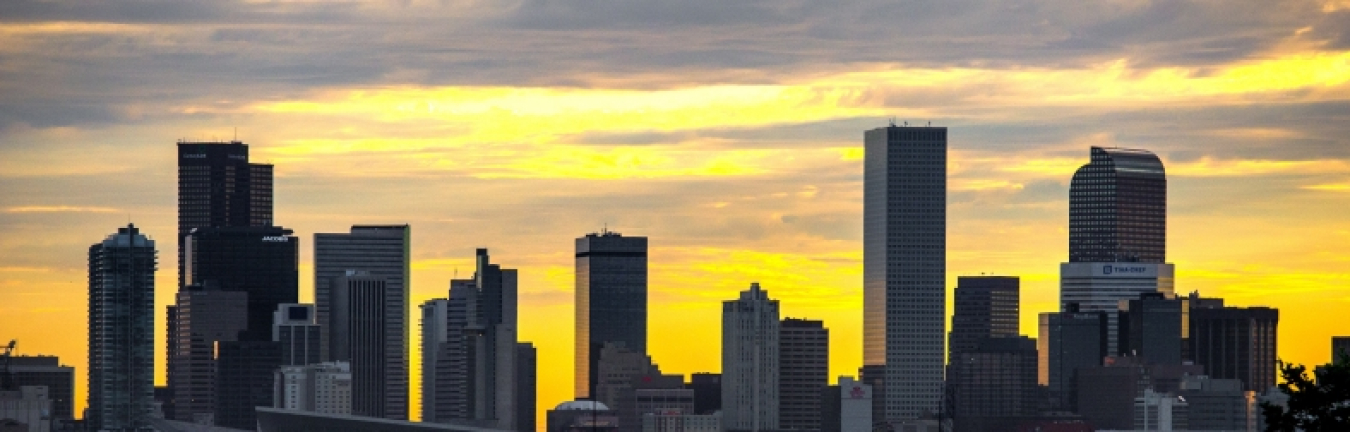 Photo of a city skyline with tall office buildings, and a multicolored sky beyond.