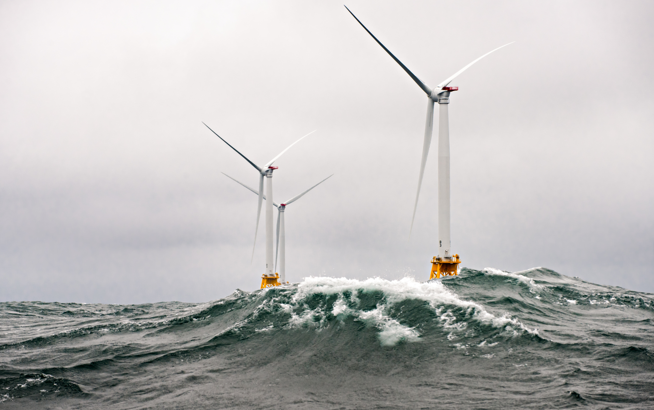 Photo of three offshore wind turbines in the open ocean, with waves cresting against them.