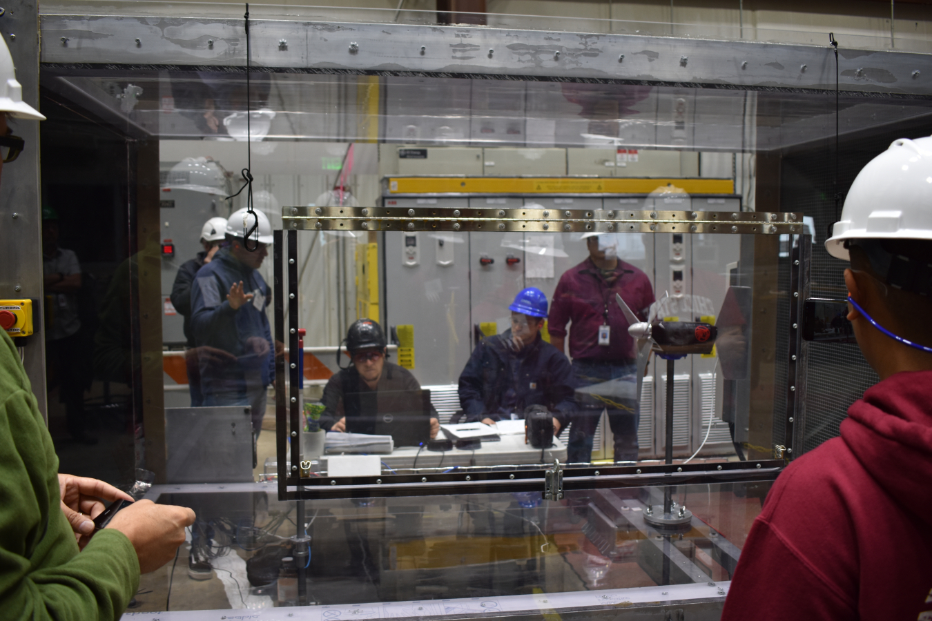 Two people stand in front of a clear wind tunnel. A turbine sits inside and a team of people running the tunnel are seated on the other side.