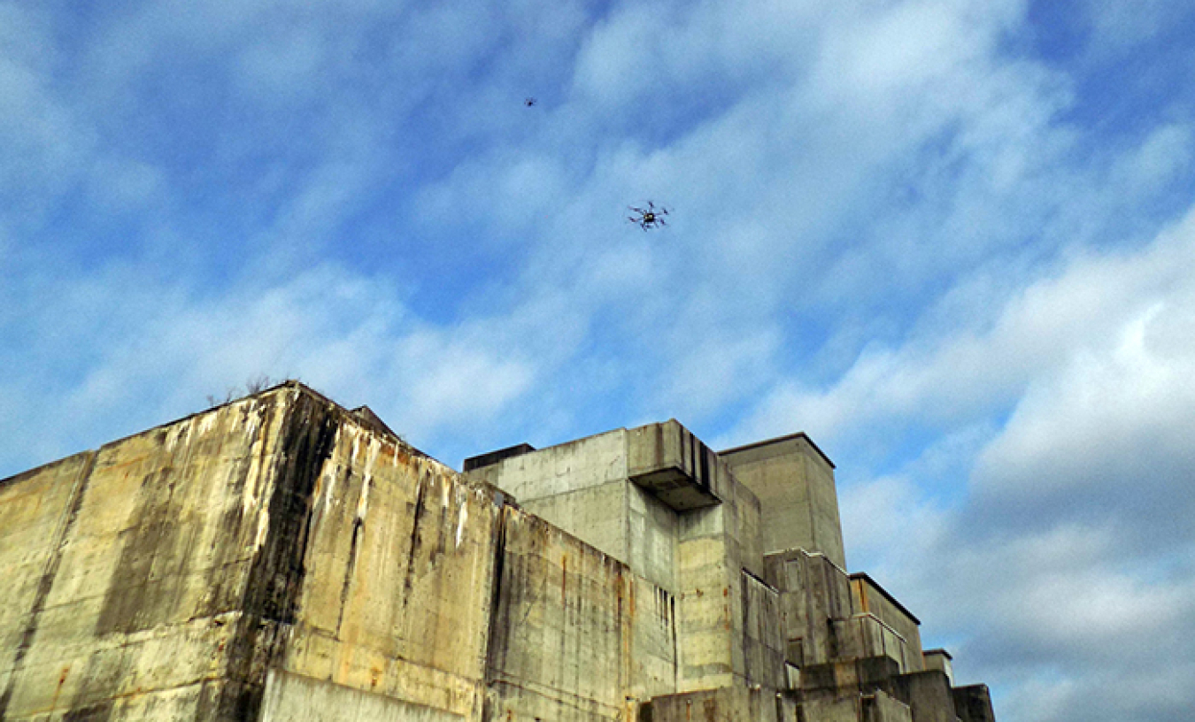 A small aerial drone with a high-resolution camera hovers above a larger drone that identifies where to spray herbicide on the rooftop of a closed reactor building.