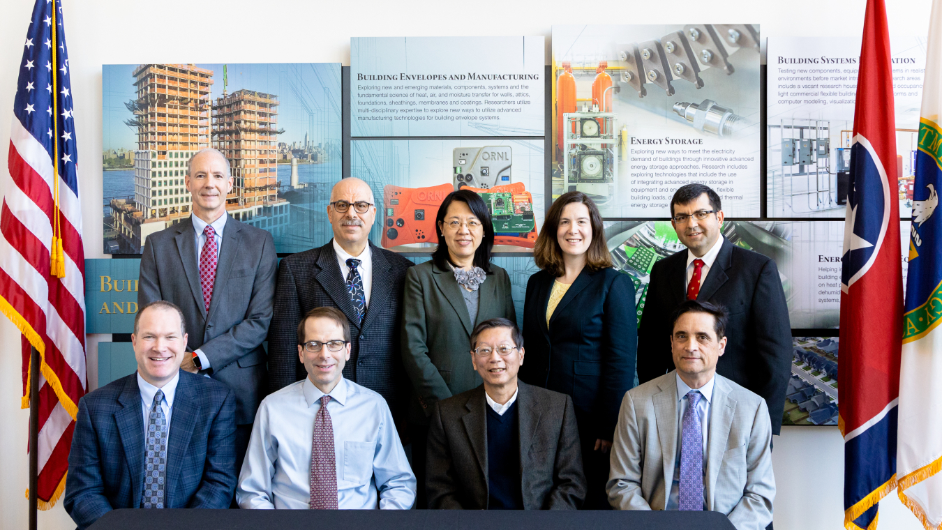 A group of nine people in business attire, lined up, facing the camera, with flags on each side.