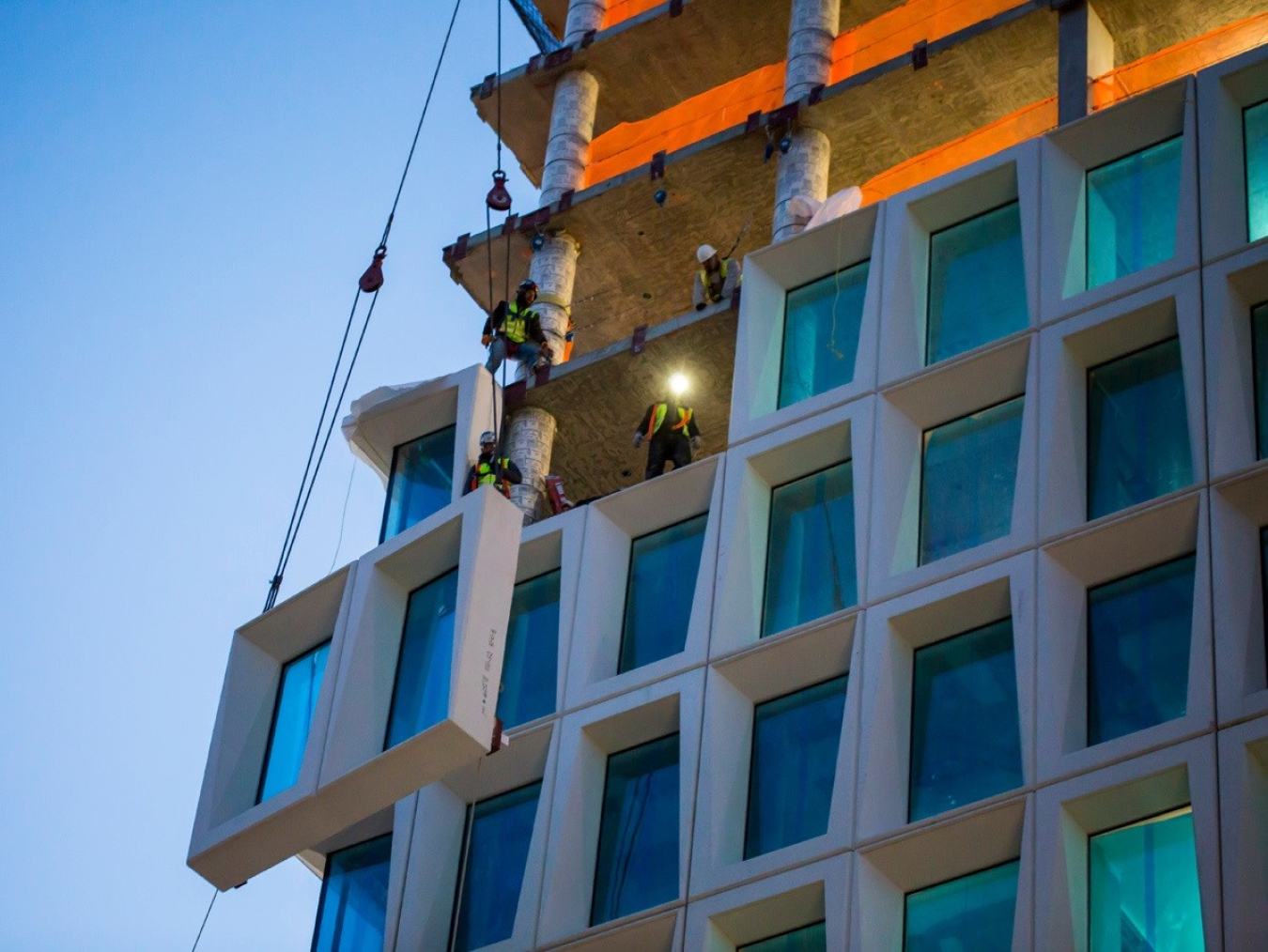 Detail of a large commercial building under construction, with workers lifting window panels into place.