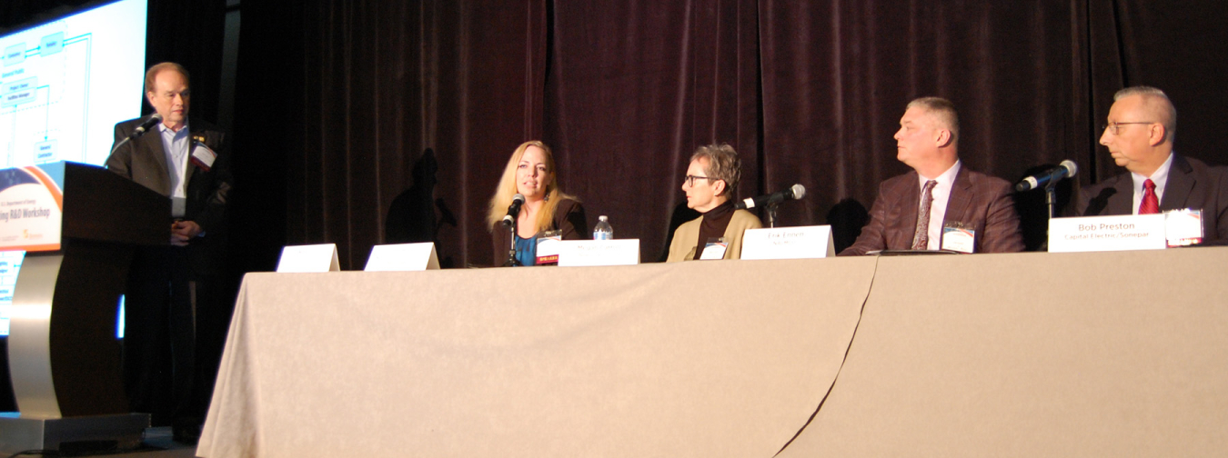 A man on a podium at a conference with a microphone, looking to his left at four additional people seated at a table also on the podium, facing the audience.