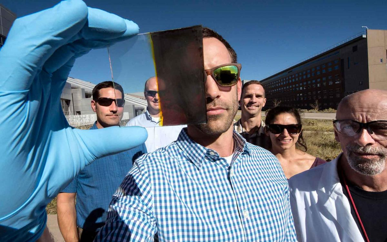 Man facing the camera holding up a small panel, with people standing behind him, in an outdoors setting.