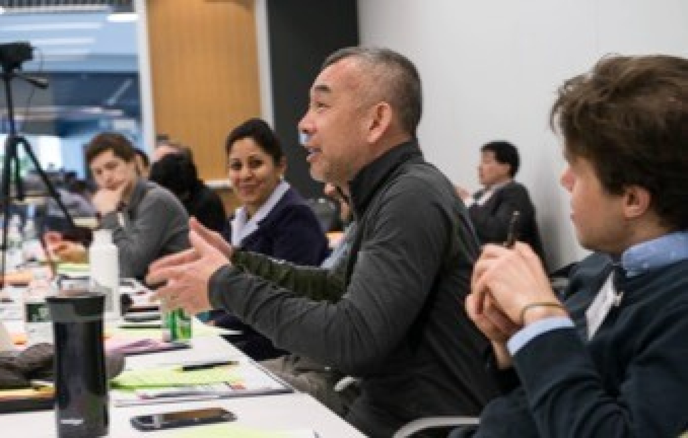 A man seated at a table, speaking, with others around him.