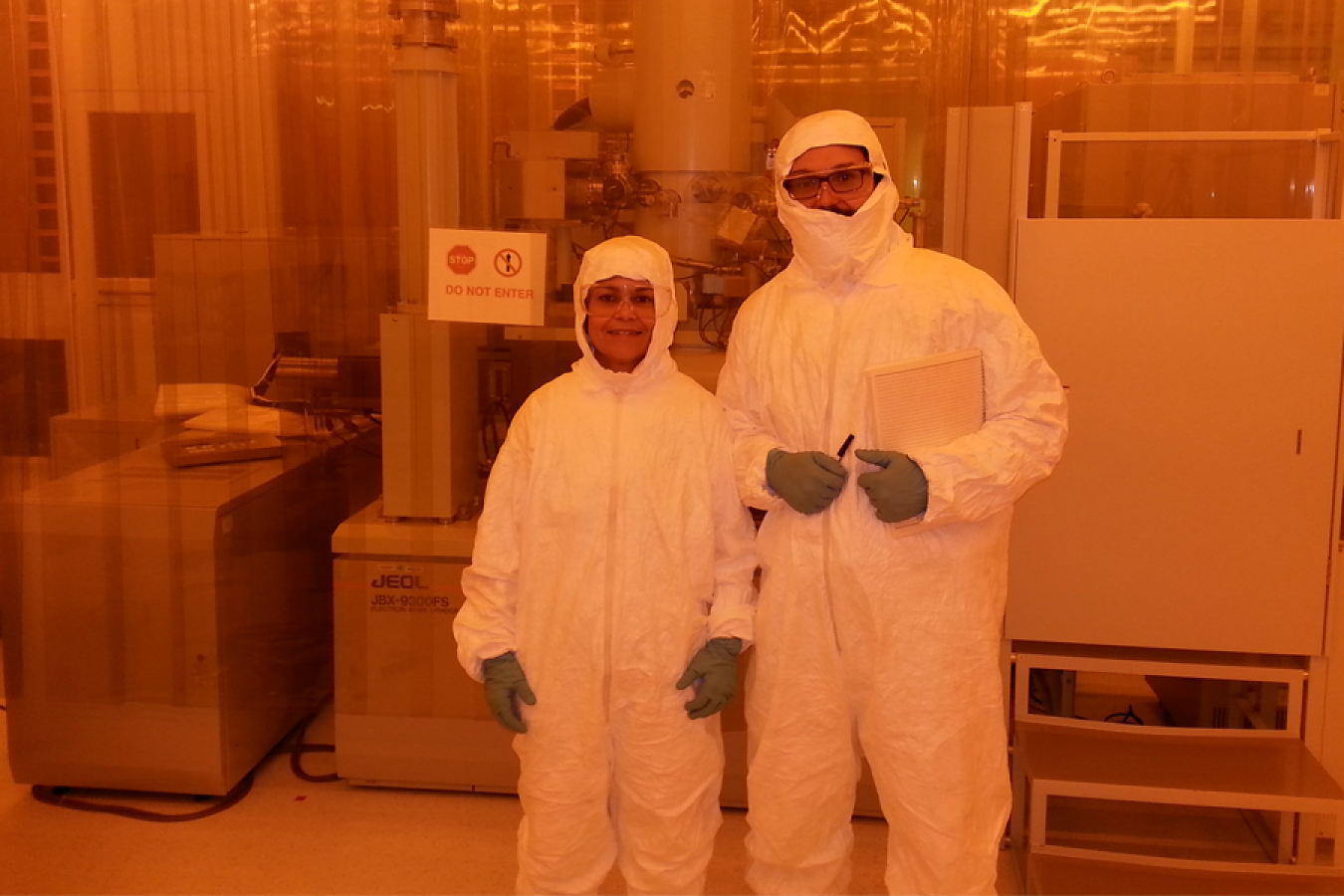 Man and woman in clean room “bunny suits” in front of machinery.