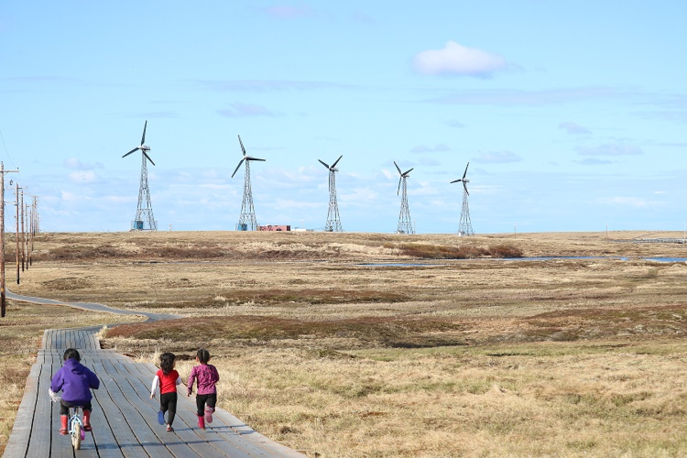 Three kids run or bike along the boardwalk in Kongiginak