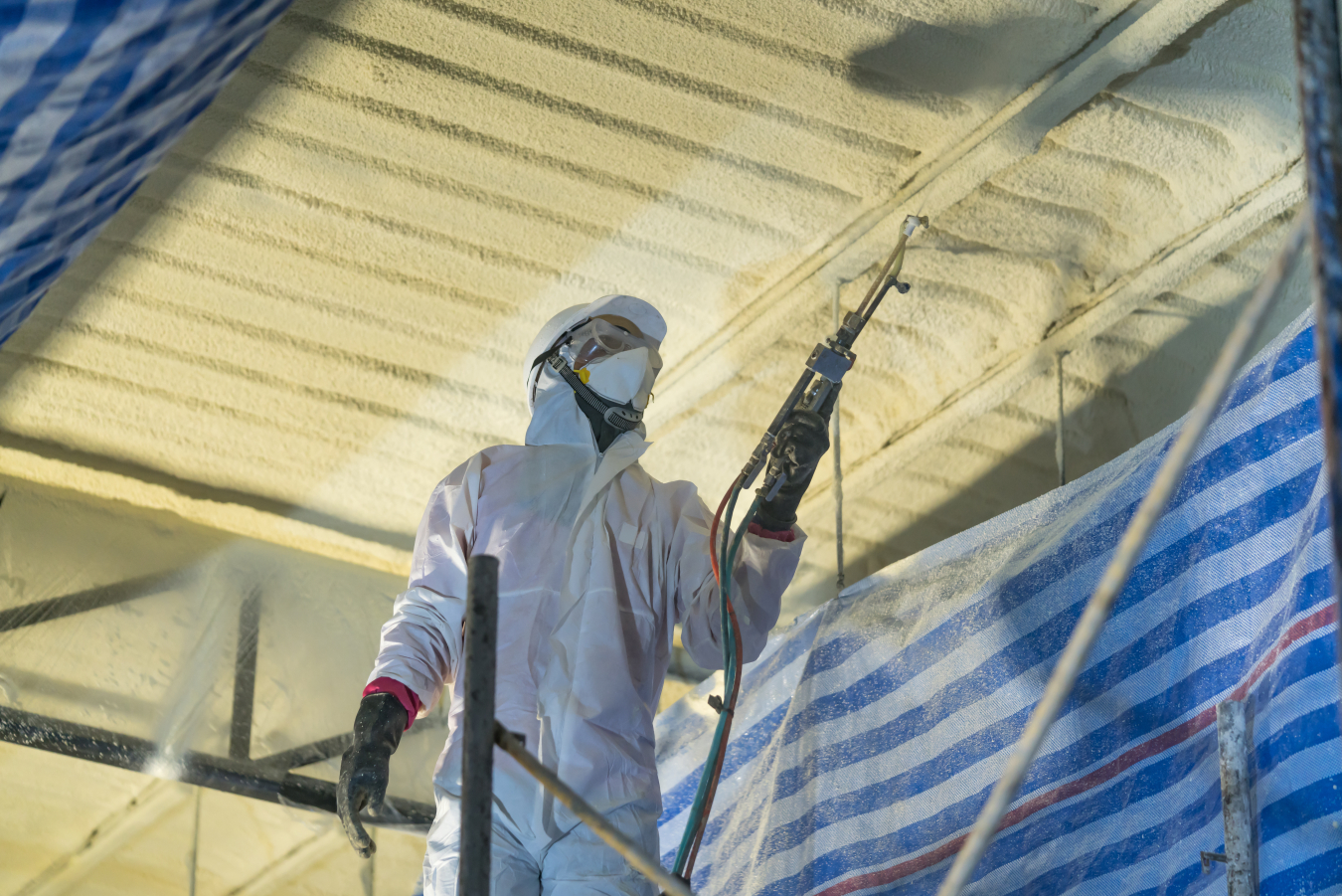 An HVAC worker wearing a protective suit and blowing in insulation for the ceiling of a building.