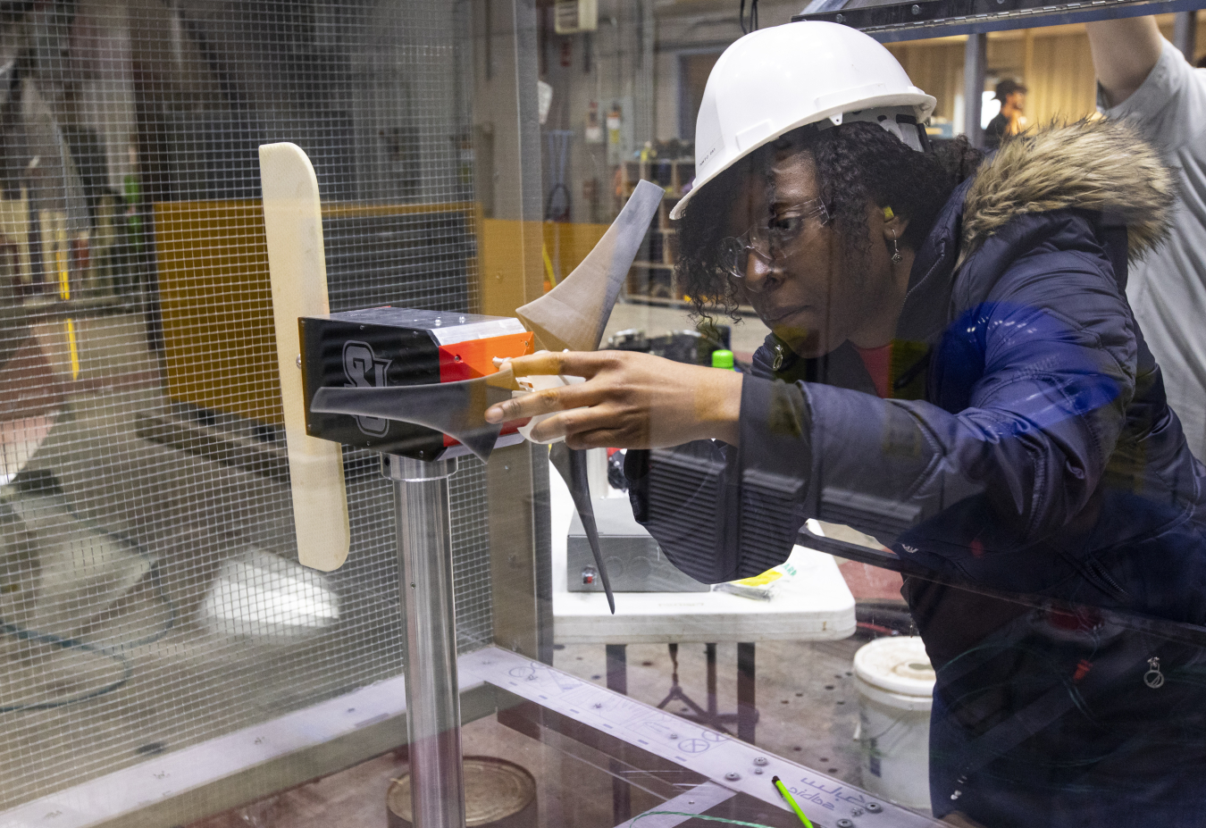 A young woman in a hard hat and safety glasses adjusts a small wind turbine in a laboratory setting.