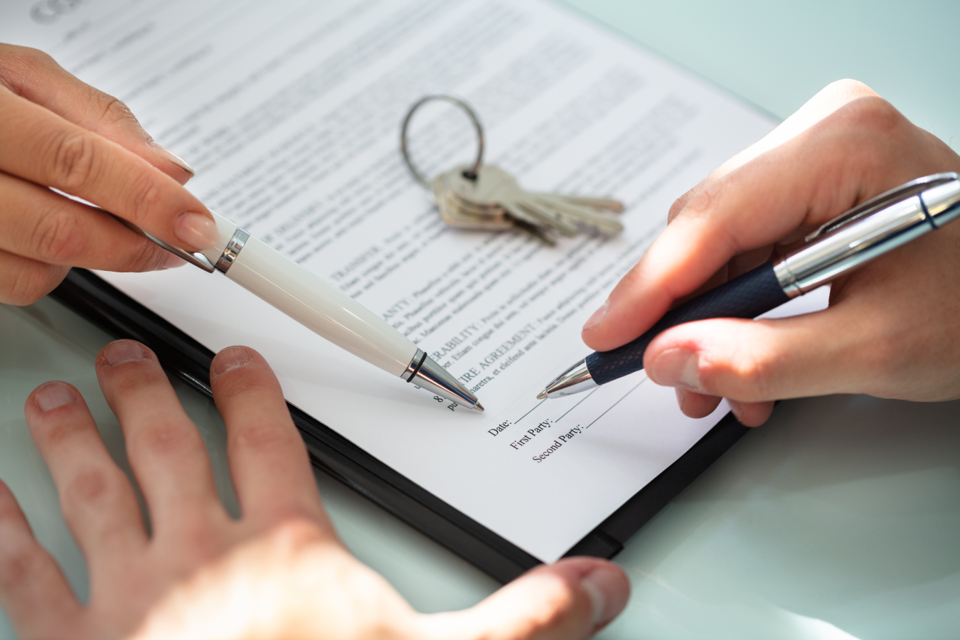 A contract on a table being signed by two people, each with a pen in hand, plus there is a set of keys sitting on top of the contract.