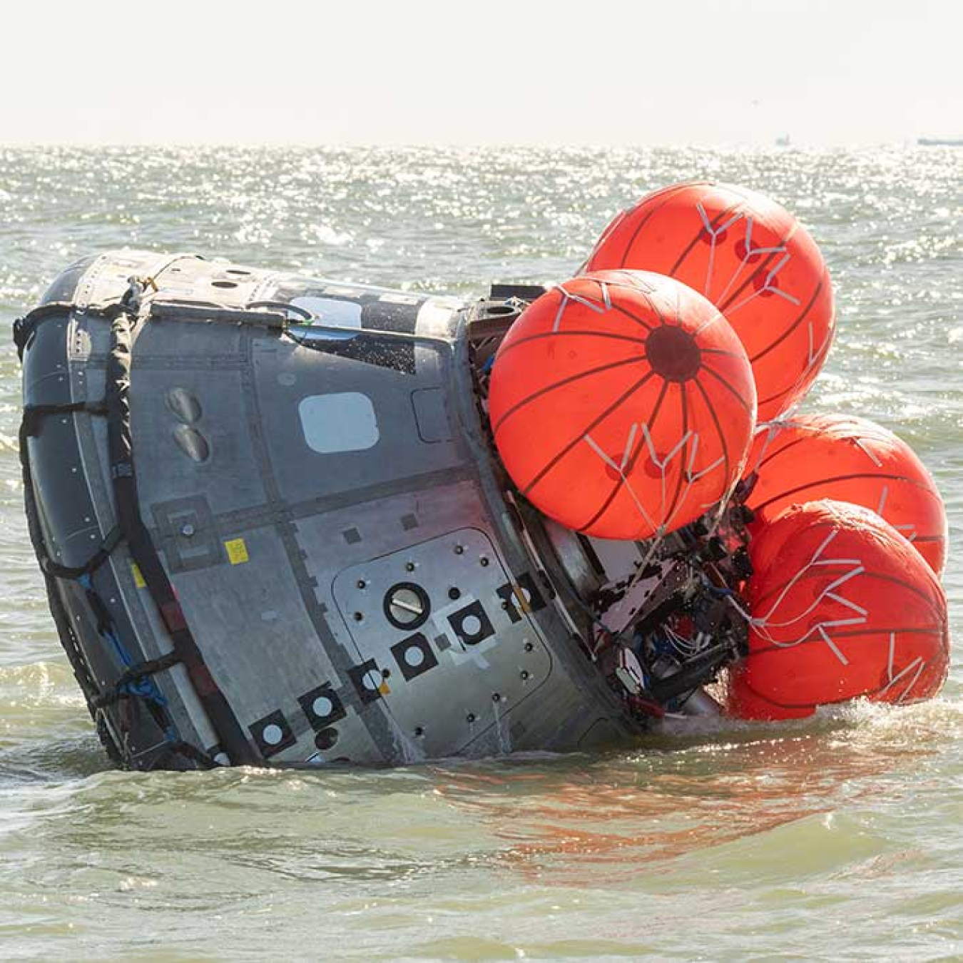 A small metallic pod floats on the ocean surface, slightly tilted on its side, as four orange balloons inflate on one end.