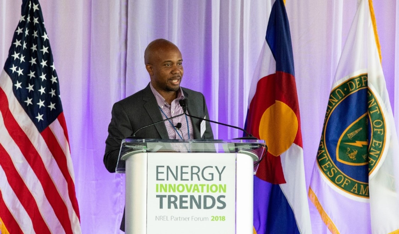 Dr. Roderick Jackson speaking at a lectern that bears the name "Energy Innovation Trends." There are U.S. and DOE flags behind him.