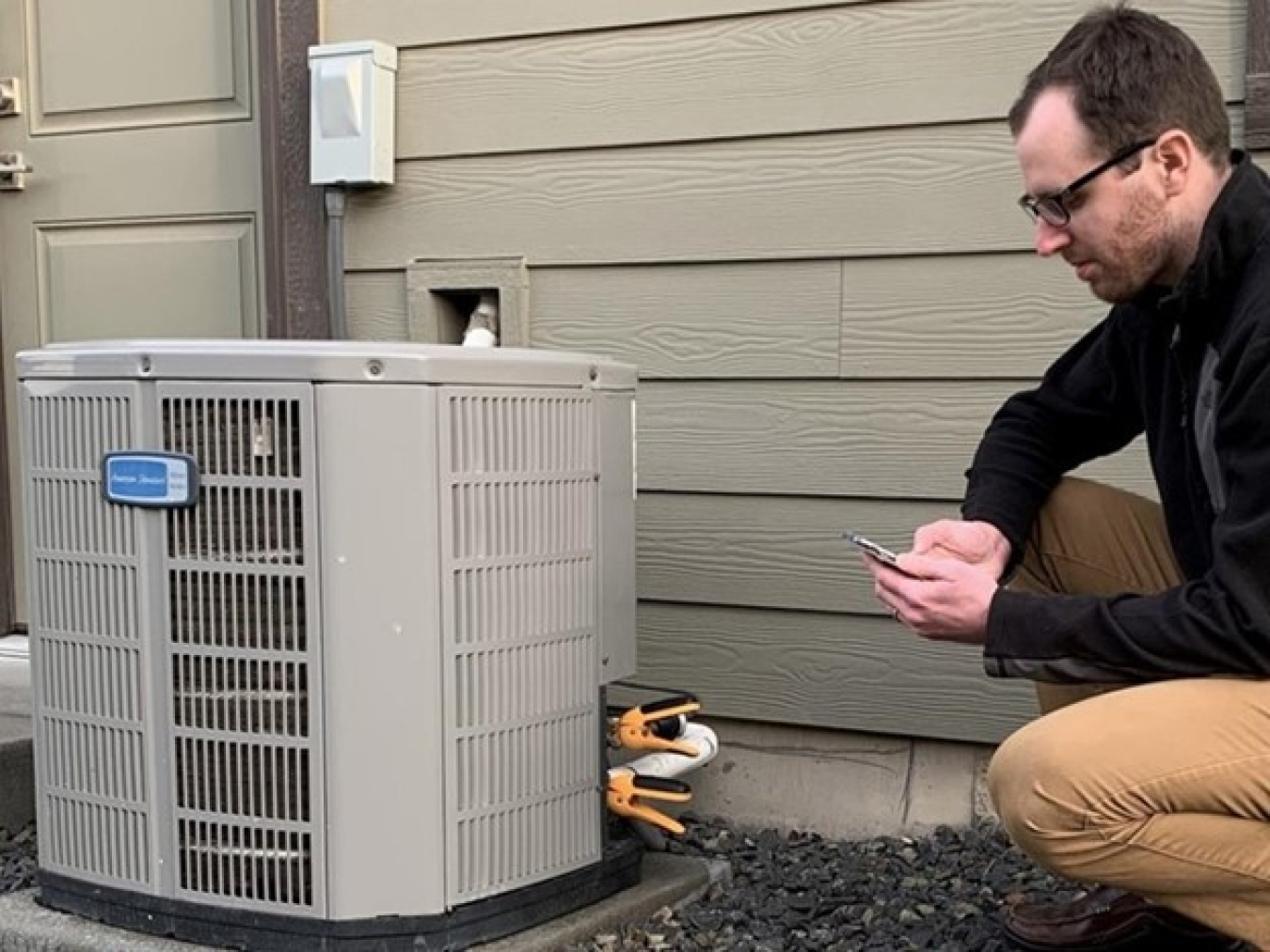 Man kneeling next to an air-conditioning unit outside a building; he's holding a device to assess it.