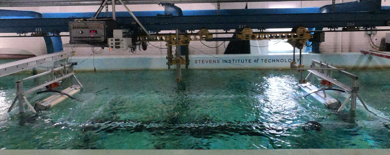 Two metal devices face each other from opposite ends of a wave tank that looks like an indoor swimming pool. Stevens Institute of Technology is written on the inside of the tank wall just above the water surface. Hanging over the water, a network of blue metal beams support a pair of electronic boxes on a track.