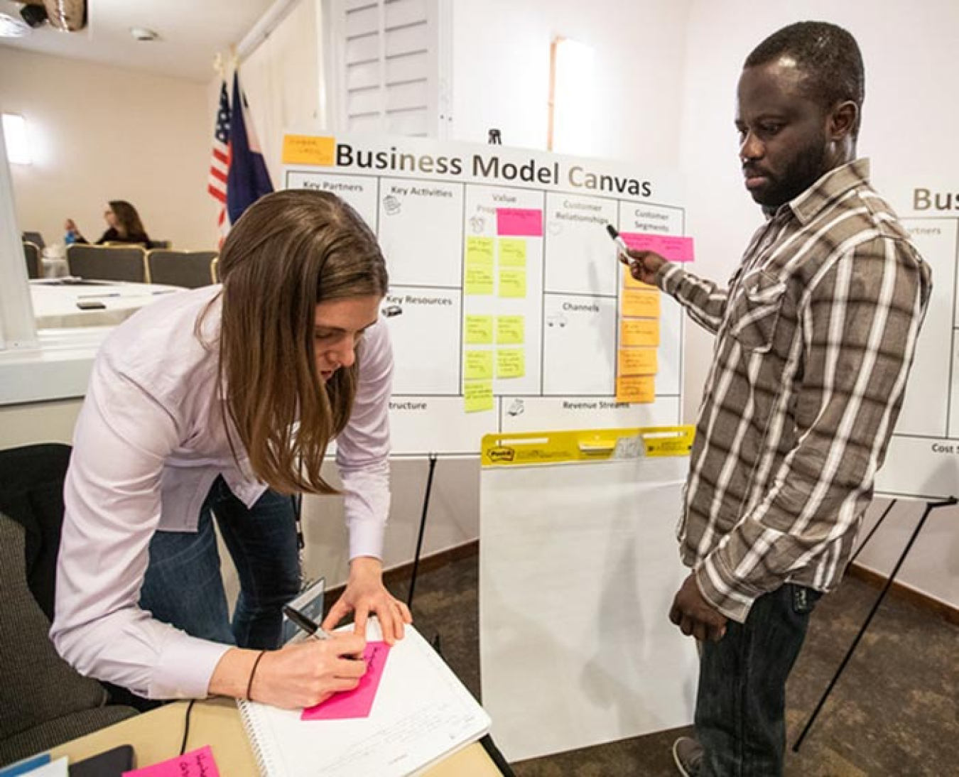 Image of a female writing on writing on a pink sticky note as a male colleague standing next to a white board looks on.