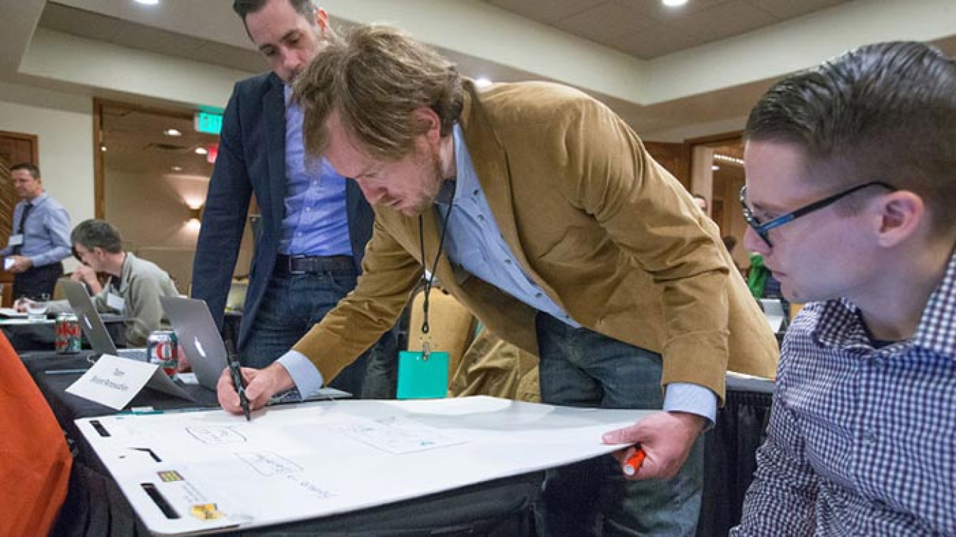 Image of three men working together at a table. The man in the middle writes on a large note pad while the other two men look on.