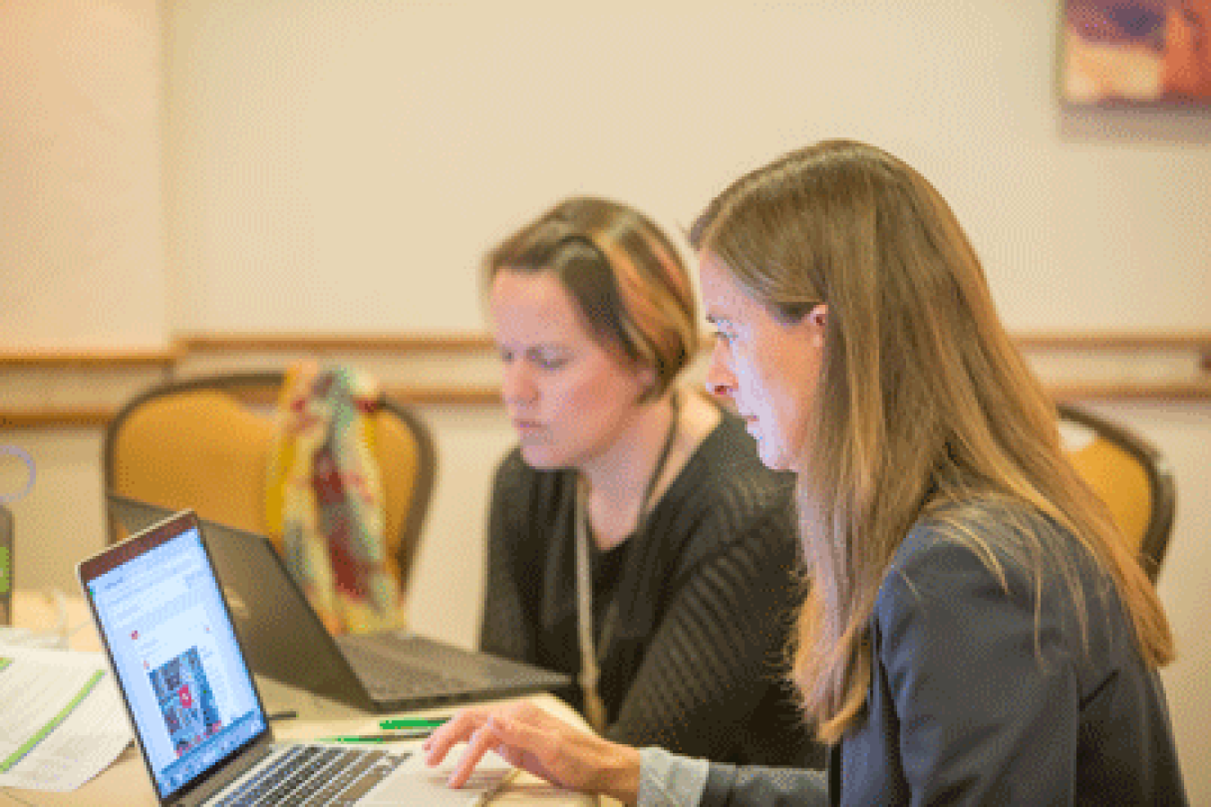 Image shows two women at a table each looking at their own laptop computers.